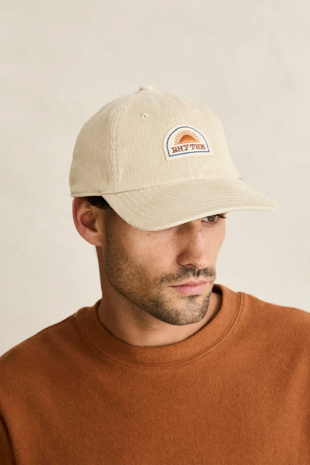 A man wearing a light beige corduroy baseball cap with a small embroidered 'Sun Town' logo on the front, paired with a brown crewneck sweater against a plain beige background.