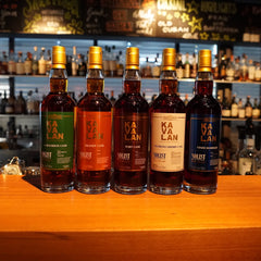 Fünf Kavalan Solist Whiskyflaschen in verschiedenen Sorten stehen nebeneinander auf einer Holztheke vor einer Bar mit vielen Flaschen im Hintergrund
