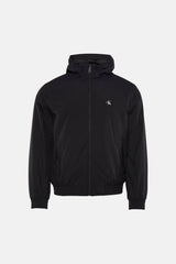 Manteau de pluie noir Calvin Klein avec capuche, porté sur une chemise noire et un jean gris, avec des baskets bleues.