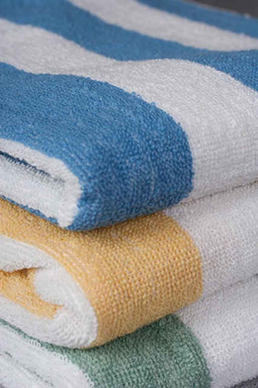 A close-up of three Ganesh Mills Cabana Stripe Beach Towels - Economy Vat Dyed, stacked with wide blue, yellow, and green stripes alternating with white. The towels have a soft, textured look.