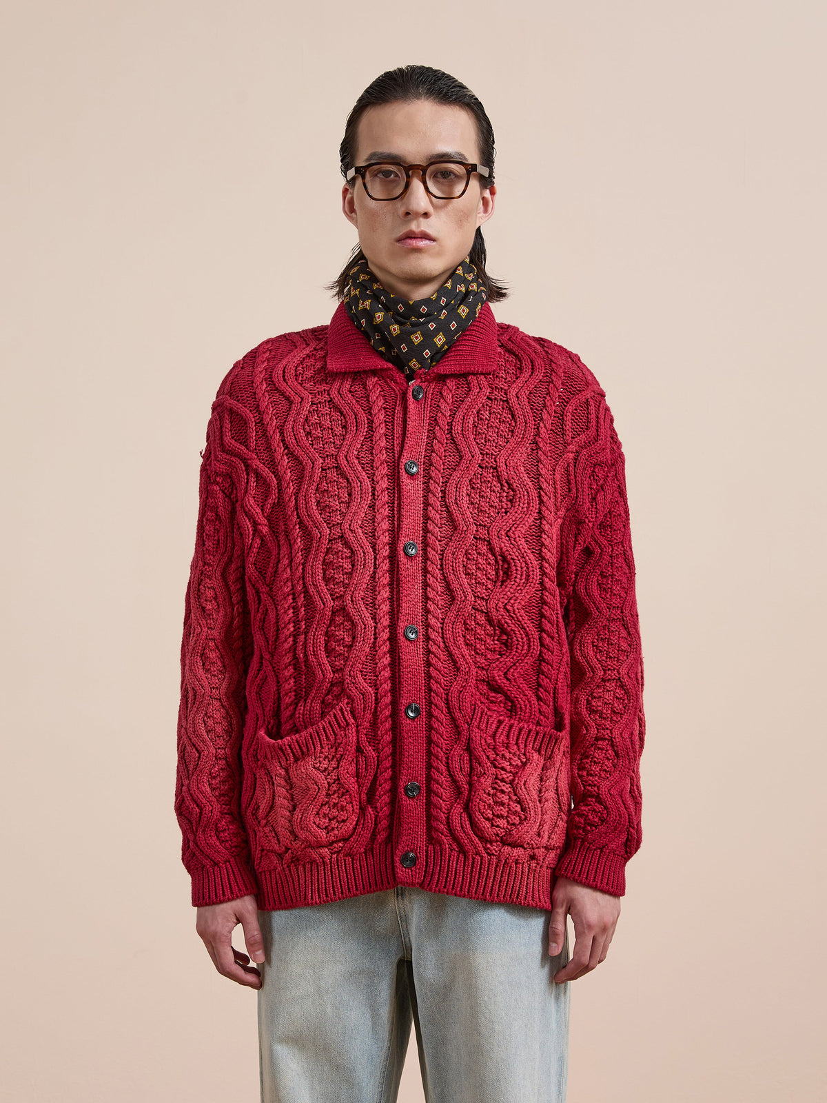 A person wears glasses, a black patterned scarf, the FOUND Cable Knit Buttoned Cardigan with pockets, and light blue jeans while standing against a plain beige background.